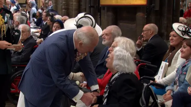King Charles shakes hands with a female veteran in a wheelchair inside Westminster Abbey. Behind him, the Queen is also speaking to veterans