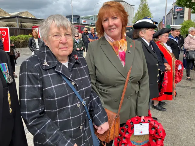 A woman wearing a grey and white patterned coat looking at the camera, A second woman is holding a wreath,