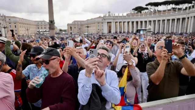 Hundreds of people in St Peter's square. Many hold up their phones or take photos or videos of the chapel rooft