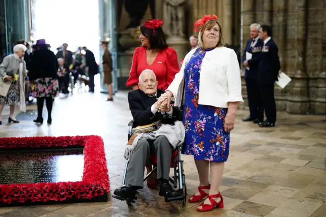 A veteran in a wheelchair holds the hand of a woman wearing a colourful dress and white cardigan as they enter into the abbey, with the entrance behind them.