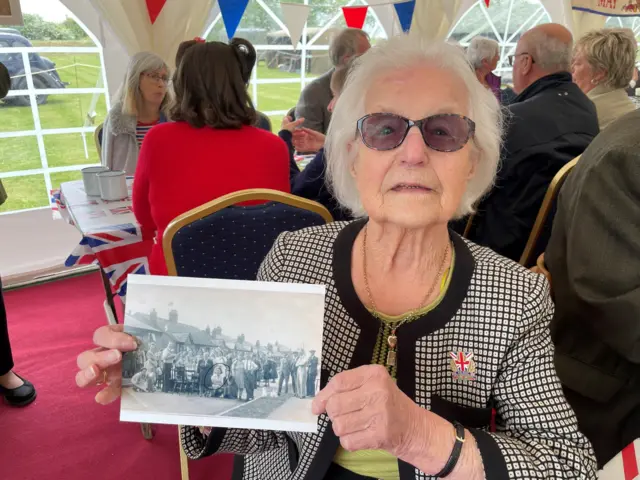 Grace Berrington who has white hair and shaded glasses holds a black and white photograph of a street party in 1945. She is sitting in a marquee with a red carpet on the floor. Other guests can be seen behind her and are sitting at long tables.