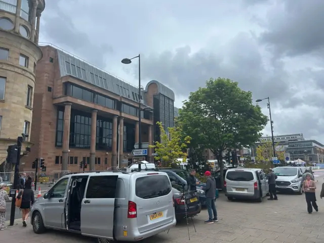 A number of silver media vans parked outside a court building with people milling around