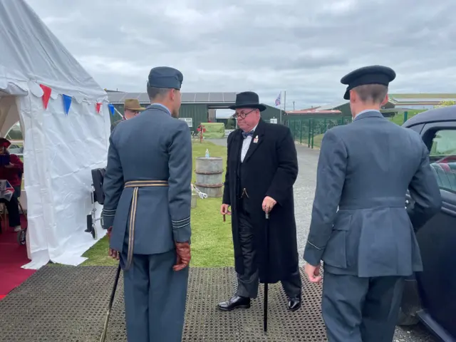 An actor dressed as Winston Churchill is met by men in RAF uniforms as he walks towards a marquee decked in bunting.