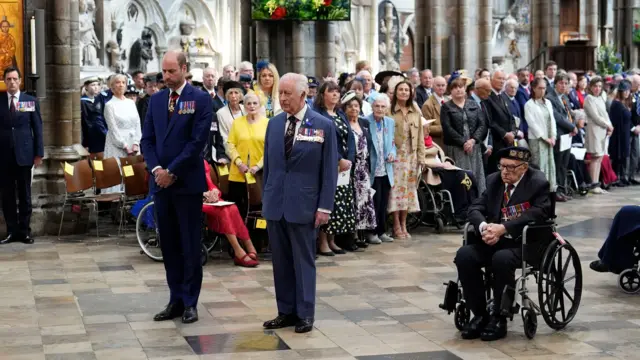 The King and Prince of Wales both stand solemnly with badges on their suits as they nod their heads in silence, with lots of attendees standing behind them and a veteran in a wheelchair to their right