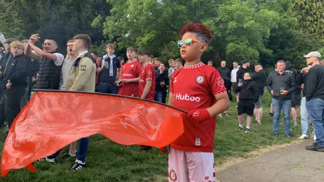 A young boy wearing a red football shirt and white shorts. He is waving a large red flag and wearing sunglasses