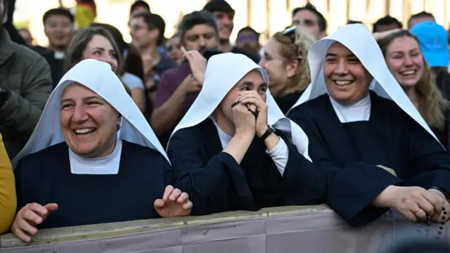 Nuns react with joy - one holds her rosary to her face, all three smile