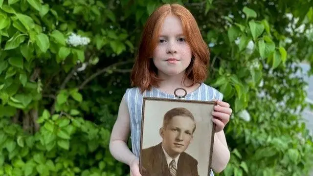 Ana holds a photo of her grandfather Albert, who served in the American navy during WW2. She is standing in front of leafy foliage.