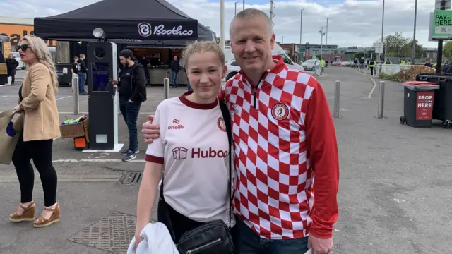 Kevin wearing a red and white Bristol City branded long-sleeved shirt. He is standing beside his daughter Megan with his arm around her. She has blonde hair in French plaits. They are both sanding by the road outside Ashton Gate Stadium.