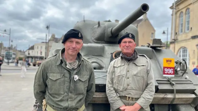 Two men in military berets and overalls standing in front of a tank, outdoors on a highstreet