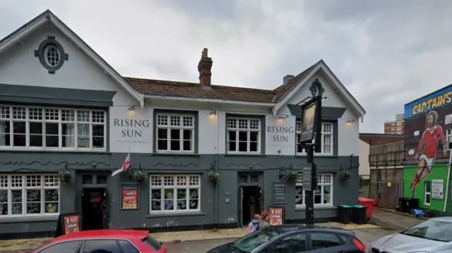 A google street view screenshot showing the outside of The Rising Sun pub. It is large and painted in two-tone grey, with a football mural next to it.