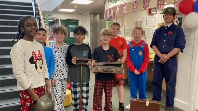 A group of seven children in pyjamas pose for a picture with a man dressed as a WW2 air warden whilst holding several objects incudlng a helmet, a spade and cardboard suitcases