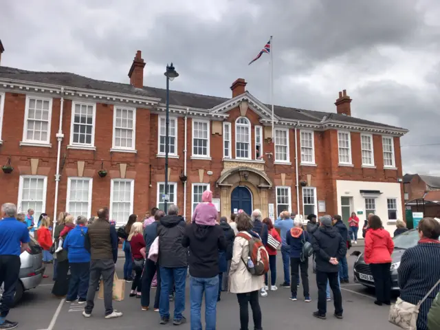 A crown gathers in front of a period red brick building with white windows. A flag pole is in the centre of the image and a red, white and blue union flag has been raised on it.
