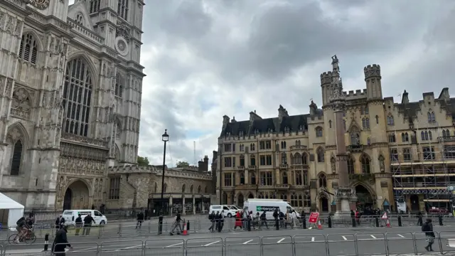 Side view of the entrance to Westminster Abbey, with metal barriers lining the empty main street as passers-by walk along