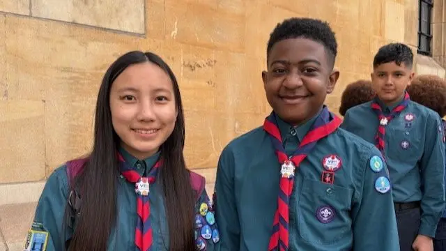 A teenage girl (L) with long brown hair wearing a green-shirt Scout uniform stands next to a teenage boy (R) wearing a matching uniform as they pose for pictures in front of a yellow stone wall