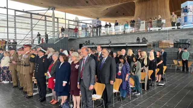 group of people, some suited and some in military uniforms, standing from their seats to observe silence.