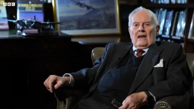 Colin Bell sits on a leather chair wearing a dark suit, dark blue vest and red and blue tie. A model of a bomber placed on a piano to his right, a bookcase and painting in a golden frame behind him