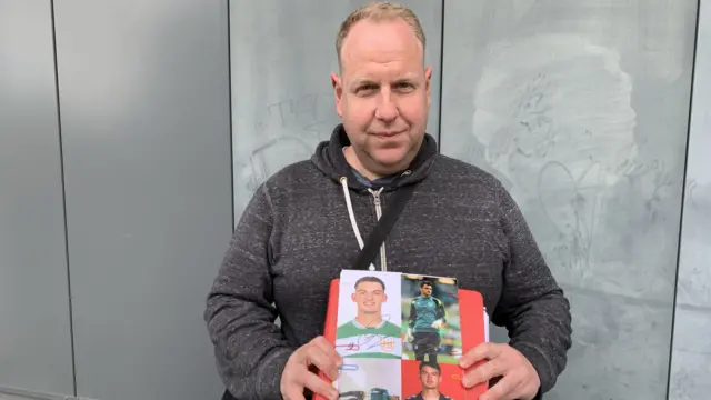 David Morgan wearing a dark grey hooded sweatshirt. He is holding up a red folder with pictures of the players alongside their signatures.