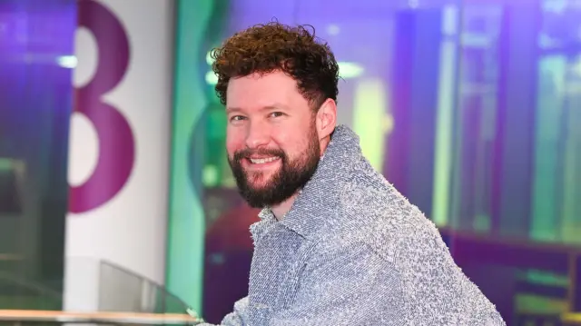 Calum Scott leans against a railing and smiles into the camera. He is wearing a blue denim jacket. He has dark, curly hair and a dark beard.