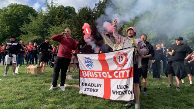 A group of men holding up a Bristol City flag and lighting off red smoke flares.