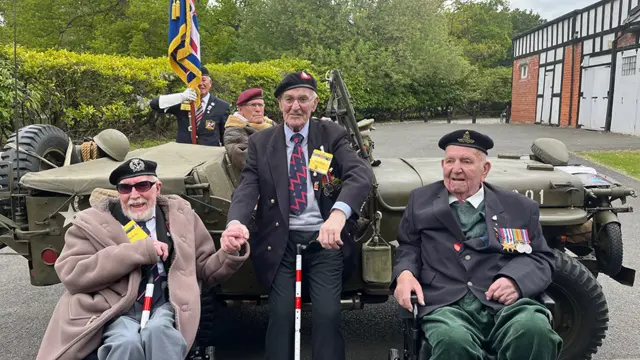 Three veterans wearing berets and medals sit in front of a World War two jeep the driver has a red beret and flying jacket and a man with blazer, medals and beret holds a military flag