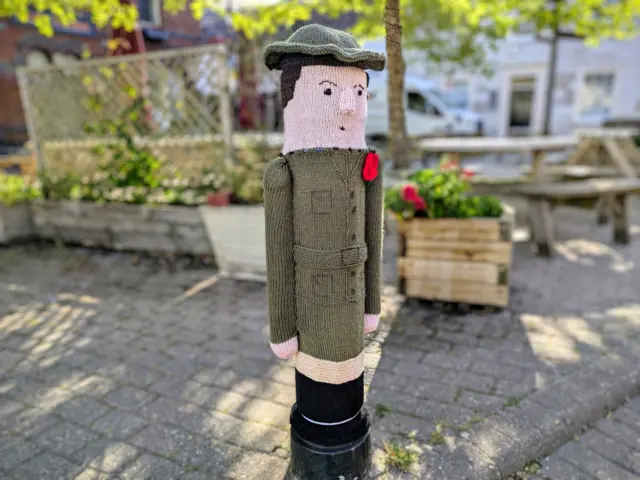 A woollen soldier in military uniform covering a bollard. He is wearing a poppy and wearing a green soldier's uniform.
