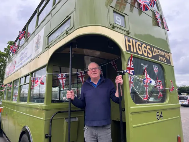 Steven Milner standing on the platform of a vintage bus. It is green and decorated with union flag bunting.