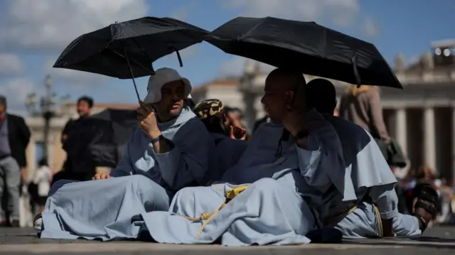 Two monks in blue robes holding black umbrella above their heads as they sit on the ground of St Peter's Square