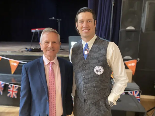 Peter Levy stands next to Vernon Kay during a live broadcast on BBC Radio 2. Peter is wearing a blue suit, white shirt and pink tie. Vernon has a tweed waistcoat and matching trousers with a white shirt and blue tie.