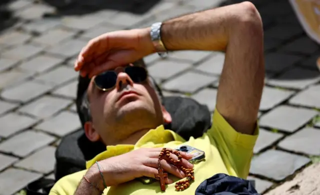 A man lying down on cobblestones, he is holding a rosary, wearing a cap and putting his face to bus forehead