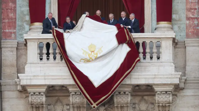 A white papal tapestry is draped over the side of the balcony.