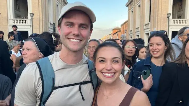 Peter Stanley and Amelia smile at the camera while stood in a crowd. Peter is wearing a beige baseball cap and top and has blue eyes. Amelia has brown eyes and brown hair tied into a ponytail. She is wearing a brown tank top.