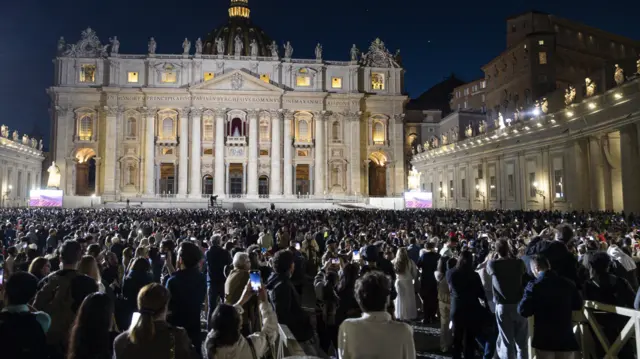A large crowd of people stands in a square surrounded by the ornate buildings of the vatican as they watch black smoke, not visible, emerge from above the Sistine Chapel.