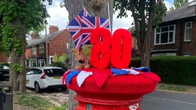 A knitted post box topper featuring a large, red 80. It also has red, white and blue knitted bunting and a brown dog.