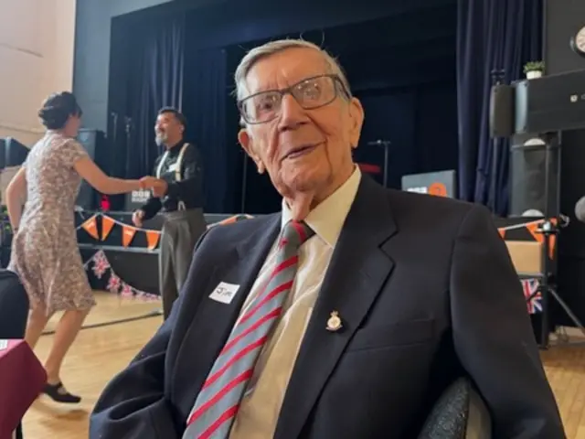 Shot of Jim Hardisty wearing a dark-coloured jacket and striped tie. There are people dancing in the background.