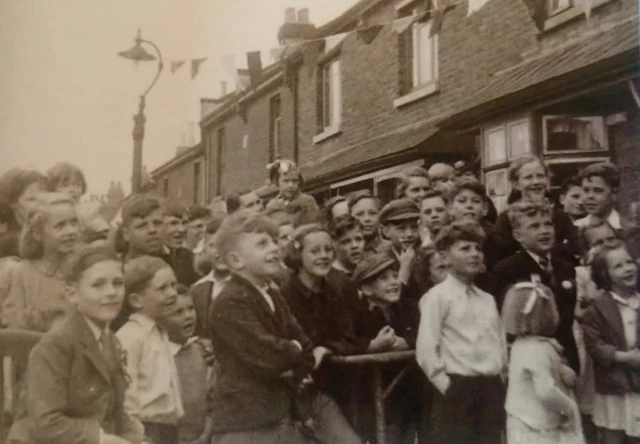 A black and white picture of children stood watching something that is out of shot at a VE Day celebration