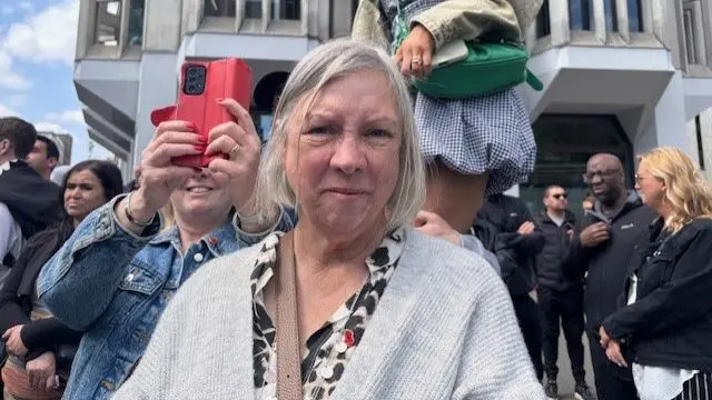 Woman, middle aged, standing in crowd outside, looking directly at camera. Wearing a white cardigan.