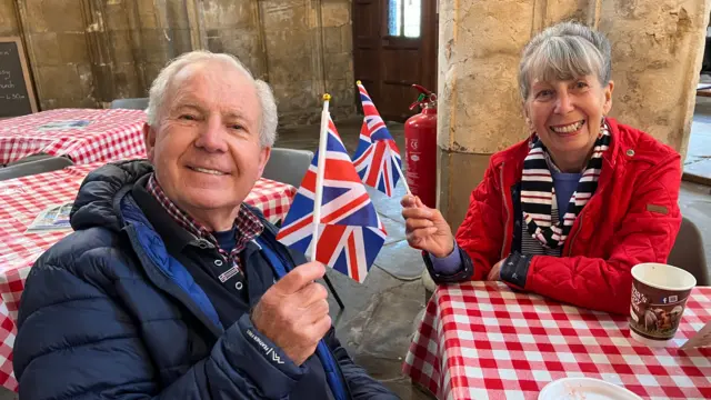 Barry Volans (L) sits at a table-cloth covered table next to his wife Gillian (R) in a blue winter jacket while holding a union flag and smiling at the camera