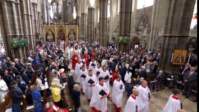 Clergy members walking down the aisle of Westminster Abbey wearing white and red robes as guests remain at their seats