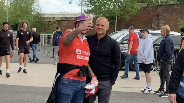 Liam Manning wearing grey trousers and a black pullover. He is outside Ashton Gate Stadium posing for a selfie with a fan, who is wearing blue jeans, an orange football top and purple bucket hat.