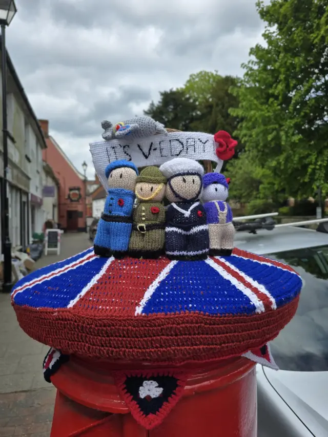 A postbox topper with a red, blue and white base marking the Union Flag and then figures, including sailors and a member of the army, on top of it.
