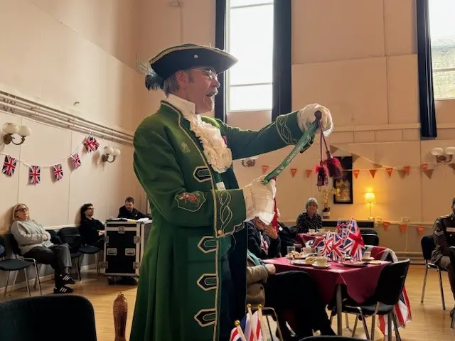 A man dressed as a town crier in a green coat with a plumed hat reads from a scroll