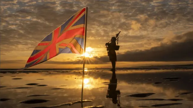National Piper Louise Marshall plays on portobello beach with a Union flag in front of him at dawn