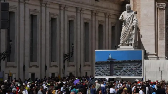 People gather near a screen displaying a live feed of the chimney on the Sistine Chapel