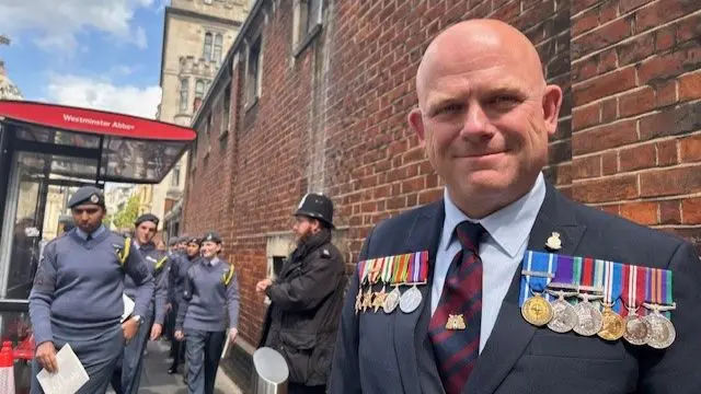 Jason Webb (bald middle-aged man) in a dark grey suit wears two sets of medals on his chest as cadets walk behind him