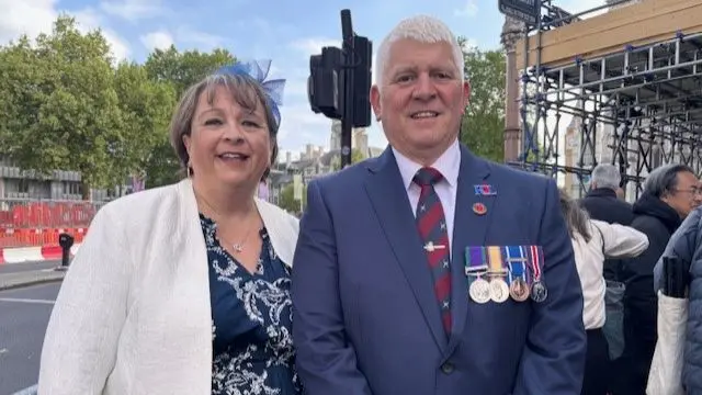Lucy Pullin (L, middle-aged woman with short brown hair) in a white blazer and blue dress stands next to her husband (R) wearing a suit with four medals on display on the left side of his chest, a queue of people to their left