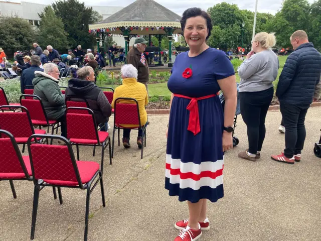 Woman with dark hair wearing a red, white and blue dress. She is standing next to a seating area and is smiling at the camera.