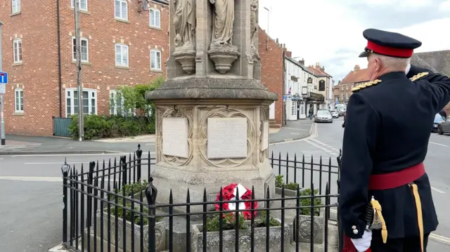 A man dressed in uniform salutes in front of the war memorial in Howden. The memorial is surrounded by black railings and features a wreath of red poppies.