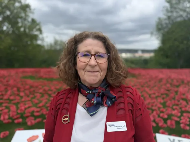 Nicky van der Drift, wearing a red jacket, white blouse and blue scarf, stands in front of a display of ceramic poppies. She has shoulder-length, curly brown hair and is wearing glasses. Trees and the skyline of Lincoln city centre are visible in the background.