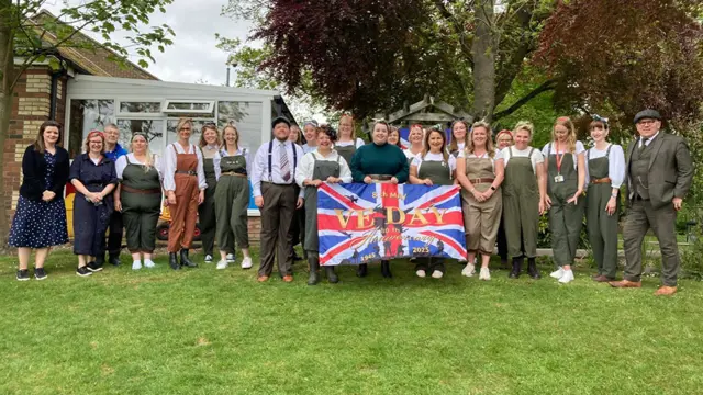 Staff at Huttoft Primary School dressed in wartime civilian clothes holding a union flag with VE Day on it