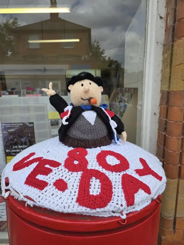 A woollen postbox topper with a red and white base with a figure of Sir Winston Churchill making a V for Victory sign.